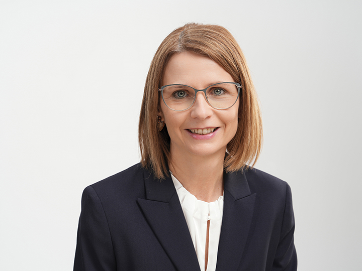 Mujer con cabello castaño claro liso hasta los hombros, que lleva gafas, un blazer azul marino y una blusa blanca, sonriendo frente a un fondo claro y sencillo.