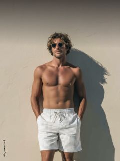 Un hombre sin camisa, con gafas de sol y pantalones cortos a rayas, está de pie frente a una pared iluminada por el sol, con las manos en los bolsillos, proyectando una sombra.
