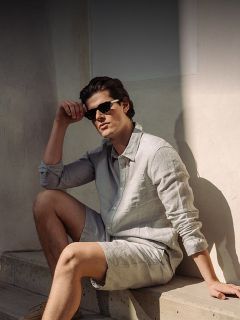 Un hombre con gafas de sol, una camisa de botones gris claro y pantalones cortos a juego está sentado en una escalera de hormigón bajo la luz del sol frente a una pared clara.