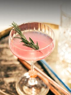 Un cóctel de color rosa, adornado con una rama de romero en una copa tipo coupe, sobre una superficie de madera rústica con vasos desenfocados al fondo.