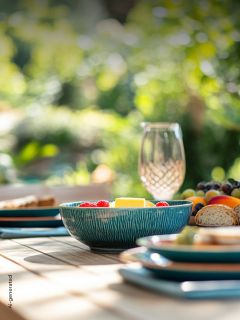 Un cuenco turquesa con fruta está sobre una mesa puesta con platos, pan y un vaso en un jardín soleado.