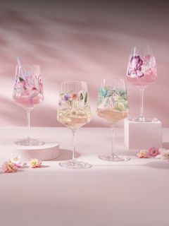 Cuatro copas de vino con coloridos diseños florales, llenas de bebidas, se encuentran sobre pedestales de colores pastel con pequeñas flores esparcidas sobre un fondo rosa.
