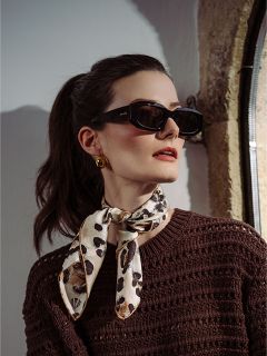 Mujer de cabello oscuro recogido en una cola de caballo, con gafas de sol negras, pendientes dorados, un suéter marrón y una bufanda con estampado de leopardo, mirando hacia un lado.