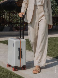 Una persona con un traje beige sale al exterior y arrastra una maleta rígida blanca con ruedas rojas y asa negra por un camino empedrado.