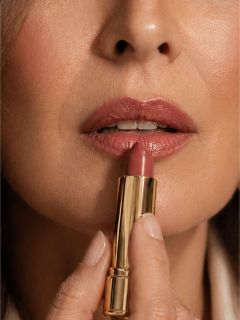 Primer plano de una mujer aplicándose lápiz labial morado en los labios con un tubo de lápiz labial dorado.