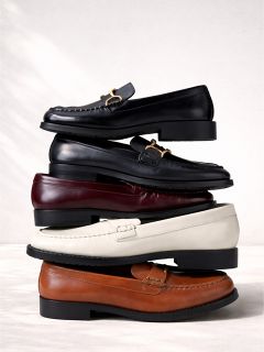 Una pila de cuatro zapatillas de cuero en negro, burdeos, blanco y marrón dispuestas verticalmente sobre un fondo claro.