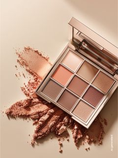 Una paleta de sombras de ojos con nueve tonalidades en colores cálidos y neutros, con un poco de polvo esparcido sobre una superficie de color beige.