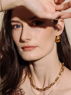 Una mujer de piel clara y cabello castaño, que lleva joyas de oro, posa con su mano protegiendo parcialmente su rostro de la luz.