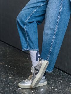 Persona con jeans azules, calcetines blancos con estampado negro y zapatillas Veja plateadas, de pie con una pierna en ángulo sobre un suelo oscuro y moteado.