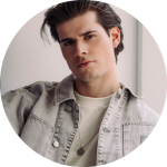 Junger Mann mit braunen Haaren, der eine helle Jeansjacke über einem weißen Hemd trägt, posiert in einem Haus und schaut in die Kamera.