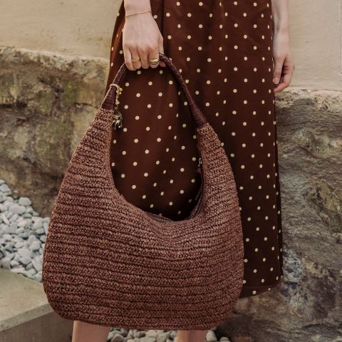 Una persona con un vestido marrón de lunares sostiene un bolso de mano grande, marrón y tejido, mientras está de pie junto a un muro de piedra y grava.