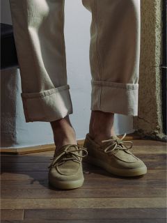 Persona con pantalones beige con puños y mocasines de ante marrón claro de pie sobre un suelo de madera cerca de la luz del sol.