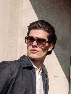 Hombre con cabello castaño peinado, gafas de sol grandes y cuadradas, chaqueta oscura y camisa clara, de pie bajo la luz del sol junto a una pared texturizada.
