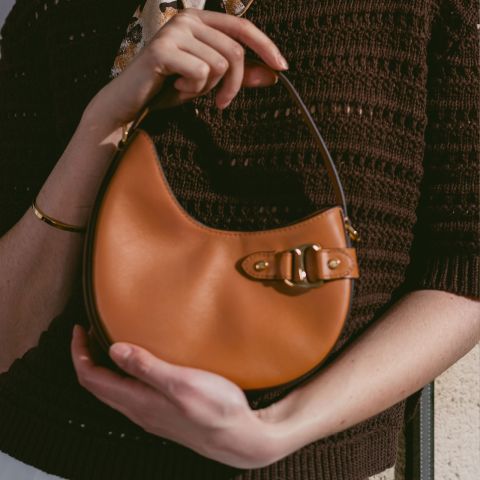 Persona con un pequeño bolso de mano de cuero marrón claro con cierre de hebilla, un top de punto marrón y una pulsera dorada.