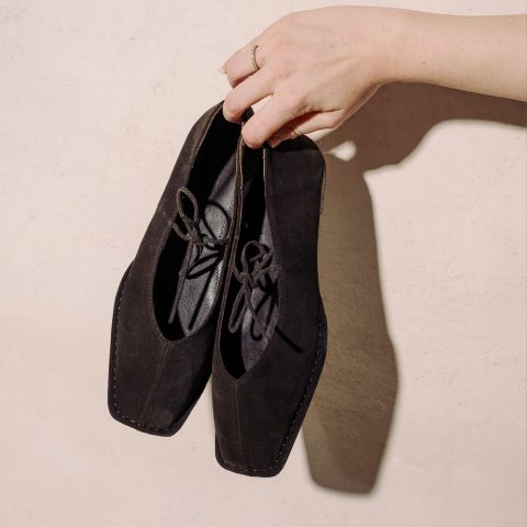 Una mano sostiene un par de zapatos de cordones de gamuza negra con punta cuadrada delante de una pared texturizada de color beige claro.