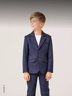 Un niño con un traje azul marino y una camisa blanca está de pie, sonriendo y mirando hacia un lado sobre un fondo claro.