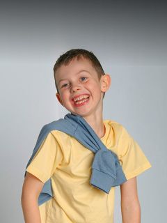 Niño sonriente con una camisa amarilla y un suéter azul sobre los hombros, de pie frente a un fondo gris sencillo.