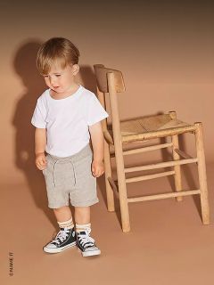 Un niño pequeño con una camiseta blanca, pantalones cortos grises y zapatillas negras está de pie junto a una silla de madera frente a un fondo beige.
