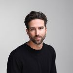 Un hombre de pelo corto castaño y barba, que lleva un jersey negro, está sentado frente a un fondo liso de color gris claro y mira a la cámara con una expresión neutra.