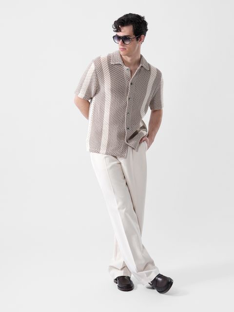Un hombre con gafas de sol, una camisa de manga corta beige con estampado y pantalones color crema está de pie con una mano en el bolsillo frente a un fondo sencillo.