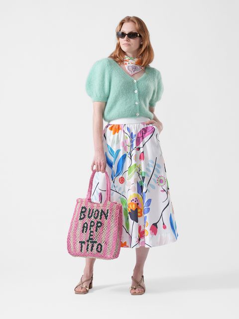 Mujer con gafas de sol que lleva una chaqueta de punto verde menta, una falda de flores, un pañuelo y sandalias, y sostiene un bolso de tela rosa con la inscripción "BUON APPETITO".