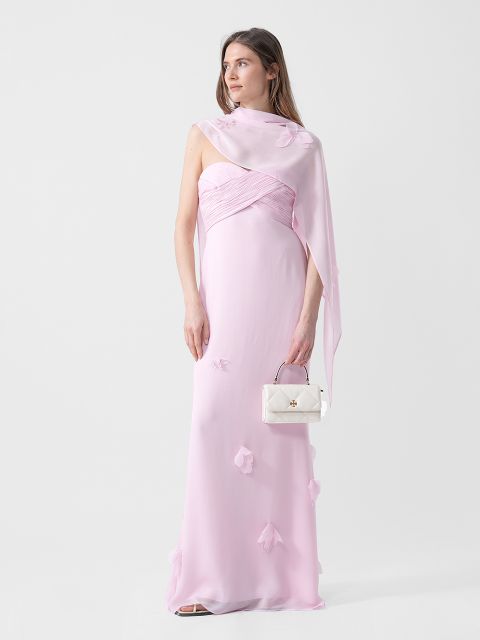 Una mujer con un vestido rosa claro sin tirantes con detalles florales sostiene un pequeño bolso de mano blanco y lleva un pañuelo transparente a juego sobre un hombro.