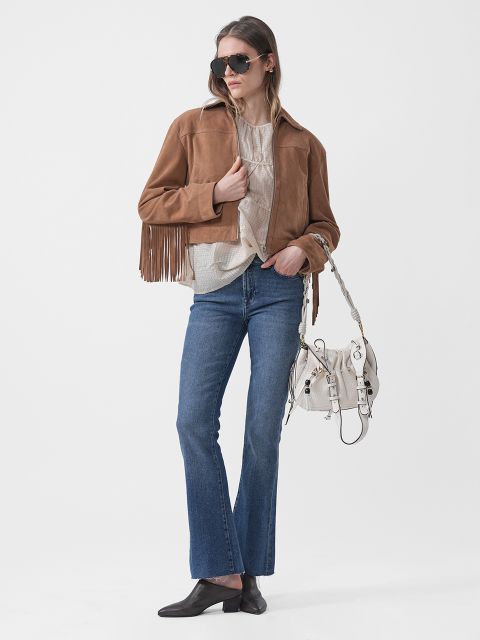 Una mujer con gafas de sol lleva una chaqueta marrón con flecos, un top beige, vaqueros azules, zapatos negros y un bolso blanco, posando frente a un fondo blanco.