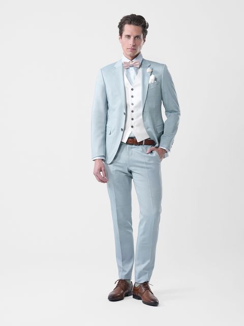 Hombre con traje azul claro, chaleco blanco, pajarita y zapatos marrones, de pie frente a un sencillo fondo blanco.