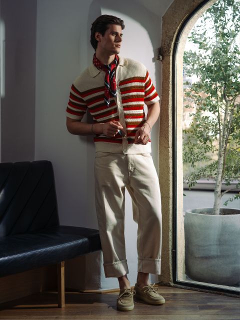 Un hombre con un polo de punto a rayas, pantalones color crema y gafas de sol posa junto a la ventana con una planta.