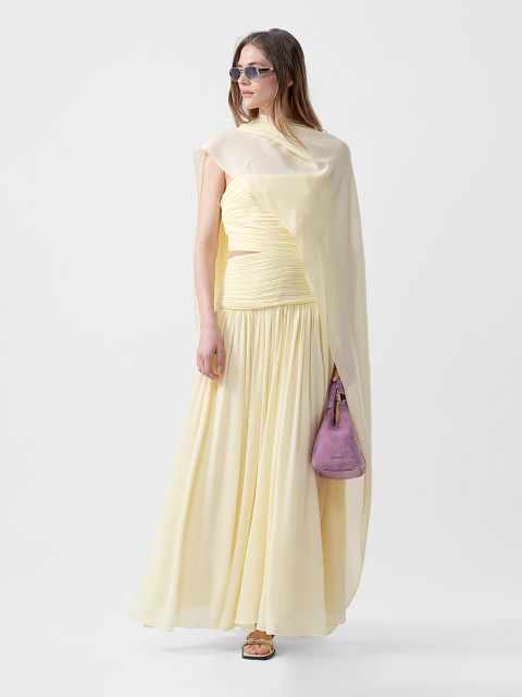 Mujer con un vestido amarillo pálido fluido con hombro drapeado, gafas de sol, sandalias y un pequeño bolso morado frente a un fondo sencillo.