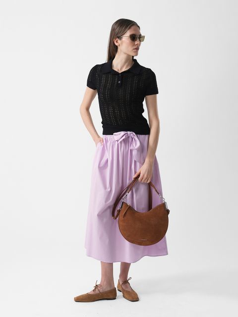 Mujer con gafas de sol que lleva una parte superior negra de manga corta, una falda de color violeta claro y zapatos marrones, sosteniendo un bolso marrón en forma de media luna frente a un fondo blanco.