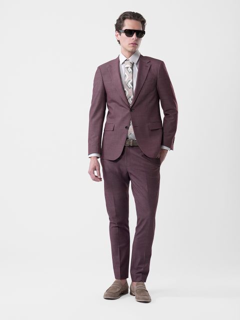 Hombre con traje morado, corbata estampada, camisa blanca, gafas de sol y mocasines beige, de pie frente a un fondo blanco liso.