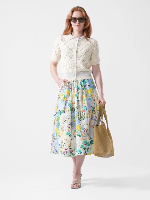 Mujer con gafas de sol, chaqueta de punto de manga corta color crema, falda estampada de flores de colores, sandalias y un bolso tote marrón claro sobre un fondo blanco.