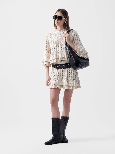 Mujer con disfraz de ganchillo color crema, botas negras, gafas de sol y un bolso negro grande de pie frente a un fondo blanco liso.
