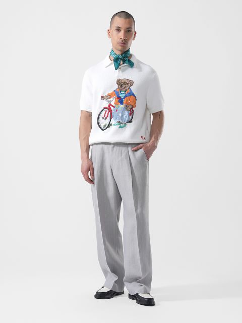 Un hombre está de pie con pantalones gris claro, una camisa blanca con un gráfico de oso, zapatos de dos tonos y una bufanda azul verdosa atada al cuello frente a un fondo blanco.