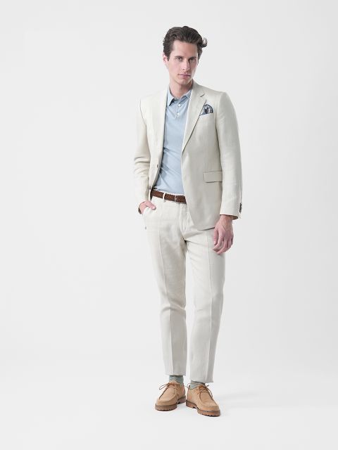 Hombre con traje color crema, camisa azul, cinturón marrón y zapatos marrones, de pie frente a un fondo blanco liso y mirando a la cámara.