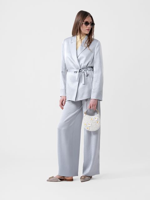 Mujer con un traje gris claro con blazer con cinturón, camisa amarilla, gafas de sol, un pequeño bolso de mano blanco y zapatos planos estampados.