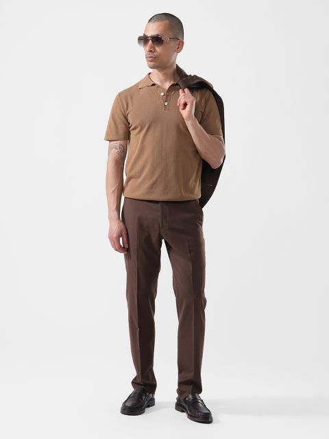 Un hombre está de pie frente a un fondo blanco, lleva gafas de sol, un polo marrón, un pantalón marrón oscuro y sostiene una chaqueta oscura sobre el hombro.
