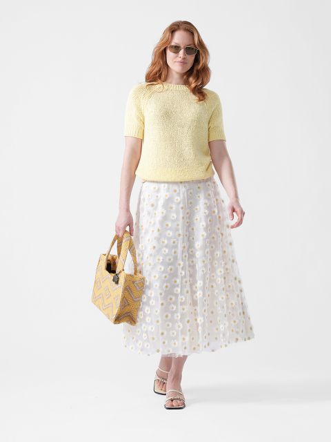 Mujer con gafas de sol, con top amarillo, falda blanca con estampado de margaritas y sandalias, sosteniendo un bolso de mano con estampado amarillo, sobre un fondo blanco.