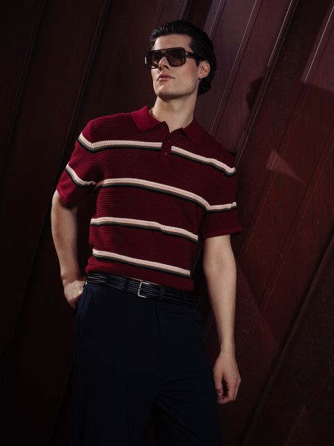 Un hombre con gafas de sol y un polo de rayas rojas posa con confianza con una mano en el bolsillo ante un fondo oscuro de paneles de madera.