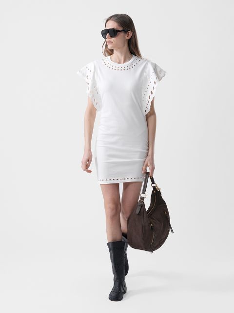 Mujer con un vestido blanco de mangas fruncidas, gafas de sol negras, botas negras y un bolso grande de color marrón, caminando frente a un fondo sencillo.