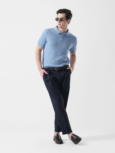 Hombre con gafas de sol, que lleva un polo de manga corta azul claro, pantalones oscuros y mocasines marrones, de pie con un pie cruzado delante del otro.