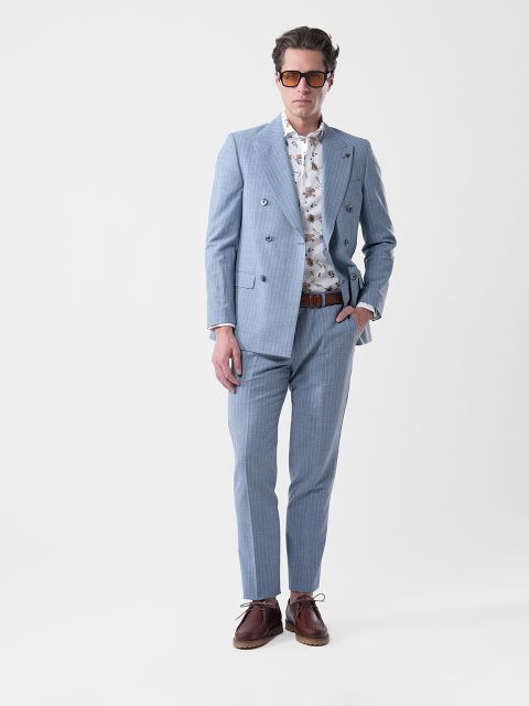 Hombre con traje azul claro, camisa blanca estampada, zapatos marrones y gafas de sol, de pie con una mano en el bolsillo frente a un fondo liso.