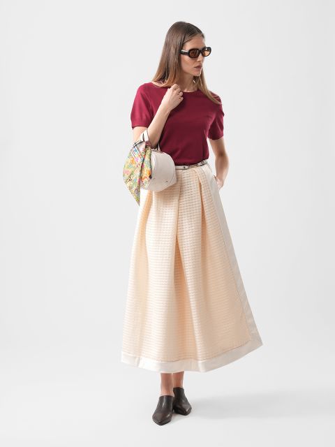 Mujer con top rojo, falda larga color crema, zapatos negros, gafas de sol y un bolso estampado en la mano, de pie frente a un fondo liso.