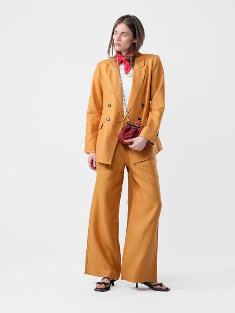 Mujer con traje amarillo mostaza, top blanco, bufanda roja, con un pequeño bolso rojo y sandalias negras, frente a un fondo blanco sencillo.