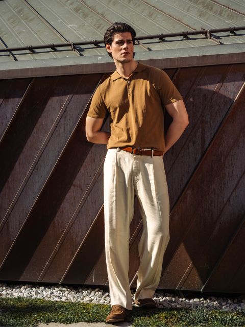 Un hombre está de pie al aire libre, lleva un polo marrón, un pantalón plisado color crema y zapatos marrones, y está apoyado en un edificio de metal a la luz del sol.