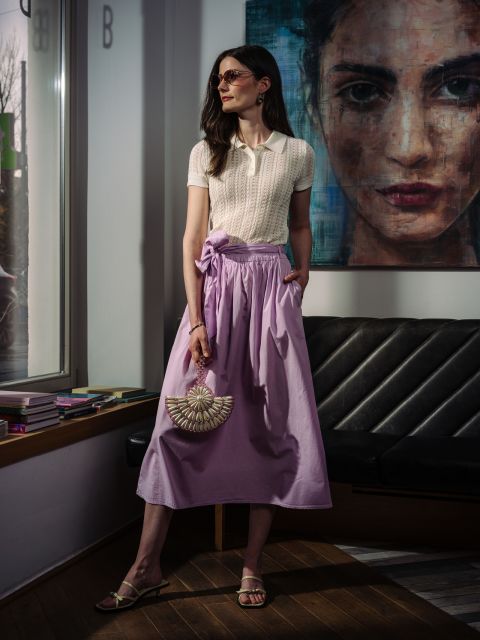 Una mujer lleva un polo color crema y una falda lavanda fluida, y sostiene un bolso de mano con forma de abanico adornado con cuentas.