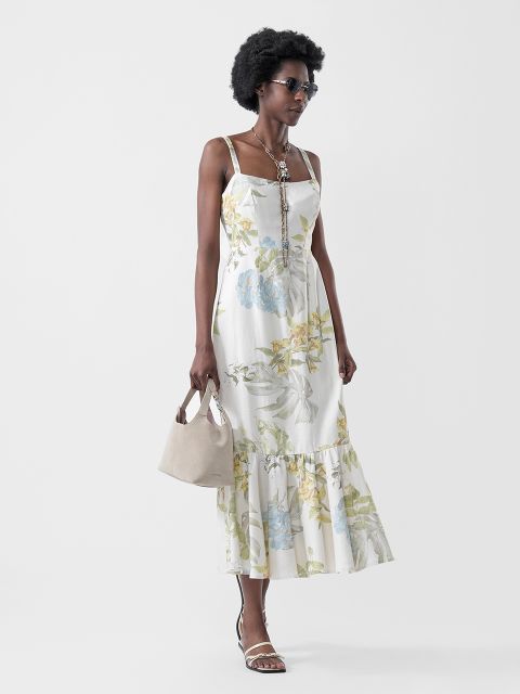 Una mujer con un vestido de verano blanco floreado, sandalias, gafas de sol y un collar, que lleva un bolso beige, posa frente a un fondo claro y sencillo.