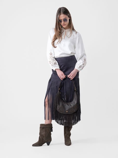 Mujer con gafas de sol, blusa blanca, falda negra con flecos, botas marrones y un bolso negro en la mano, frente a un fondo sencillo.