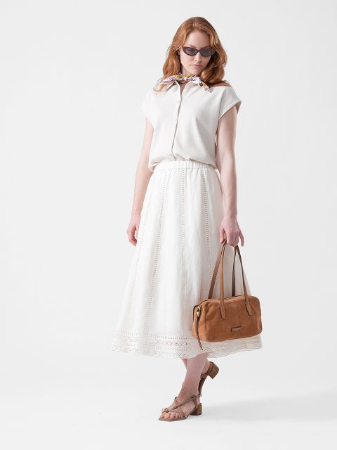 Mujer con gafas de sol, blusa blanca y falda larga blanca, que sostiene un bolso de mano marrón claro y lleva sandalias beige, de pie frente a un fondo blanco.